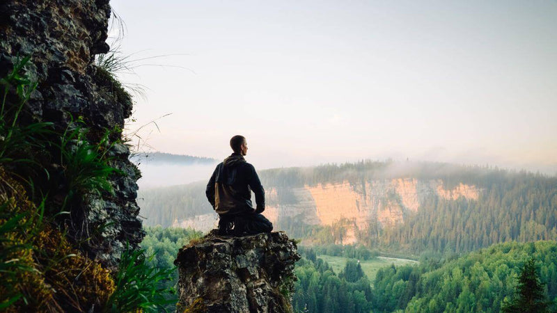 Mindfulness - An exciting path to more presence and inner peace: A person meditates on rocks overlooking nature.