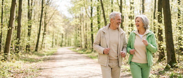 Frau und Mann um die sechzig gehen lachend einen hellen Weg durch lichten Laubwald